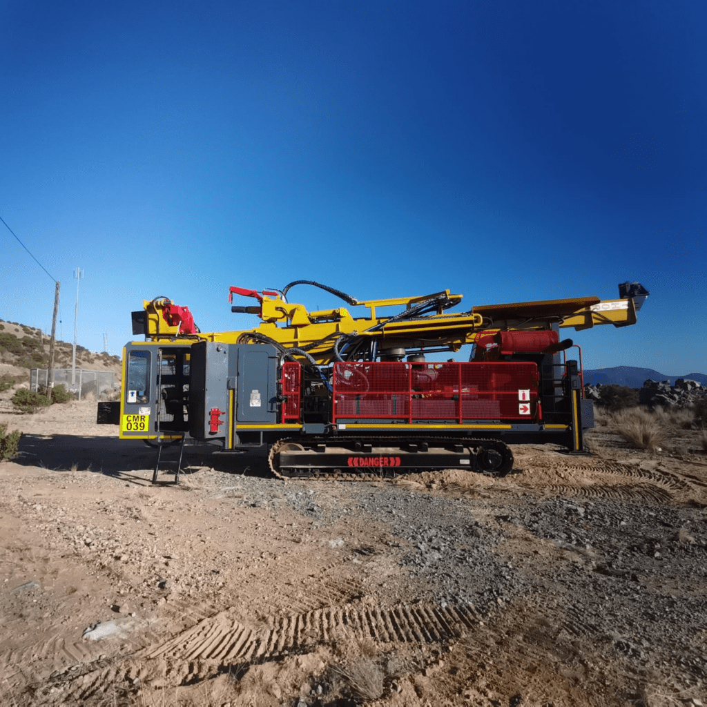 A large mining machine on a rocky outdoor landscape under a clear blue sky. The machine features a red and yellow hydraulic arm, track-mounted base, and "Danger" sign. Hills and vegetation are visible in the background.