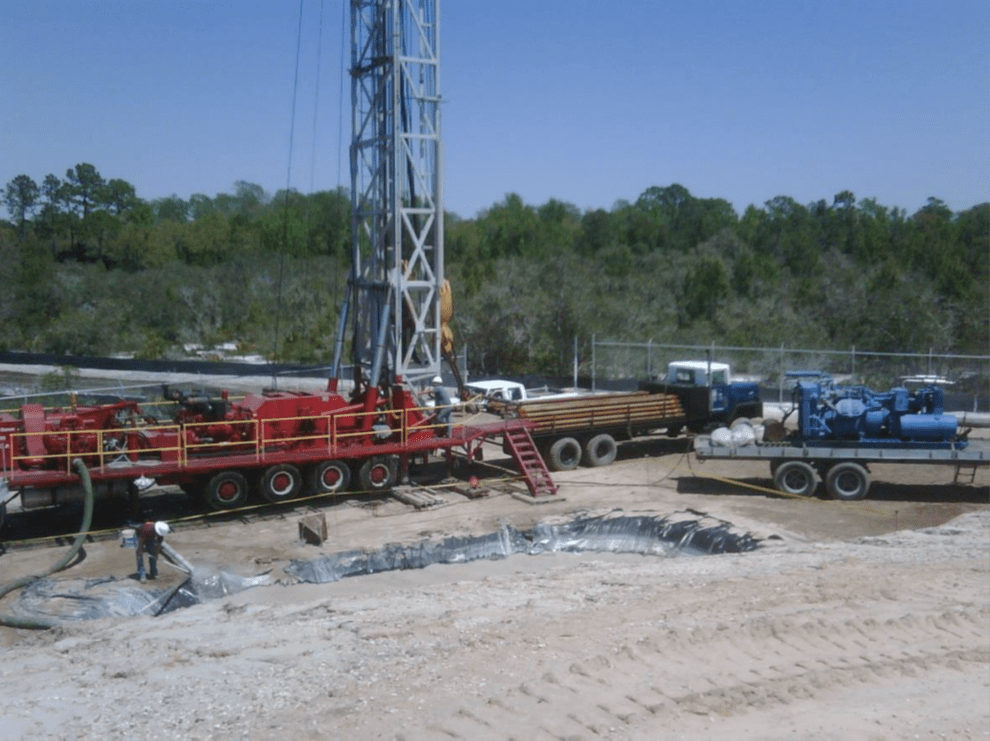 A drilling site with large machinery and vehicles, including a red truck and a blue machine on trailers. A worker in a white helmet is visible near a lined pit. Trees and a clear blue sky are in the background.