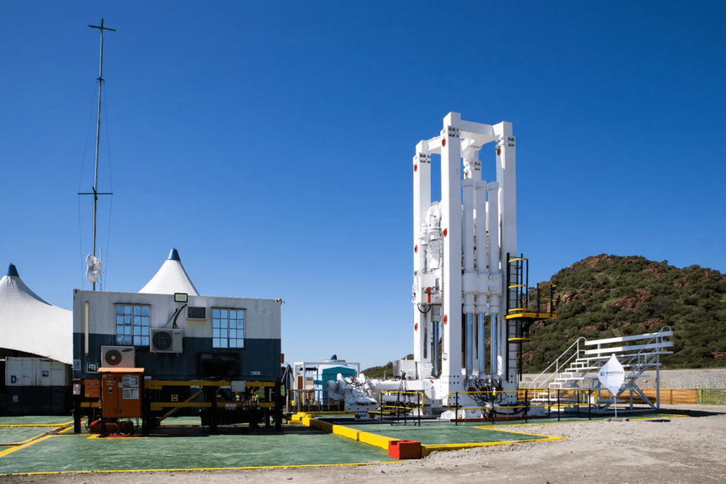 A white, vertical space launch structure stands on a prepared flat surface with a trailer and tent-like structures nearby under a clear blue sky. A hill with greenery is visible in the background.