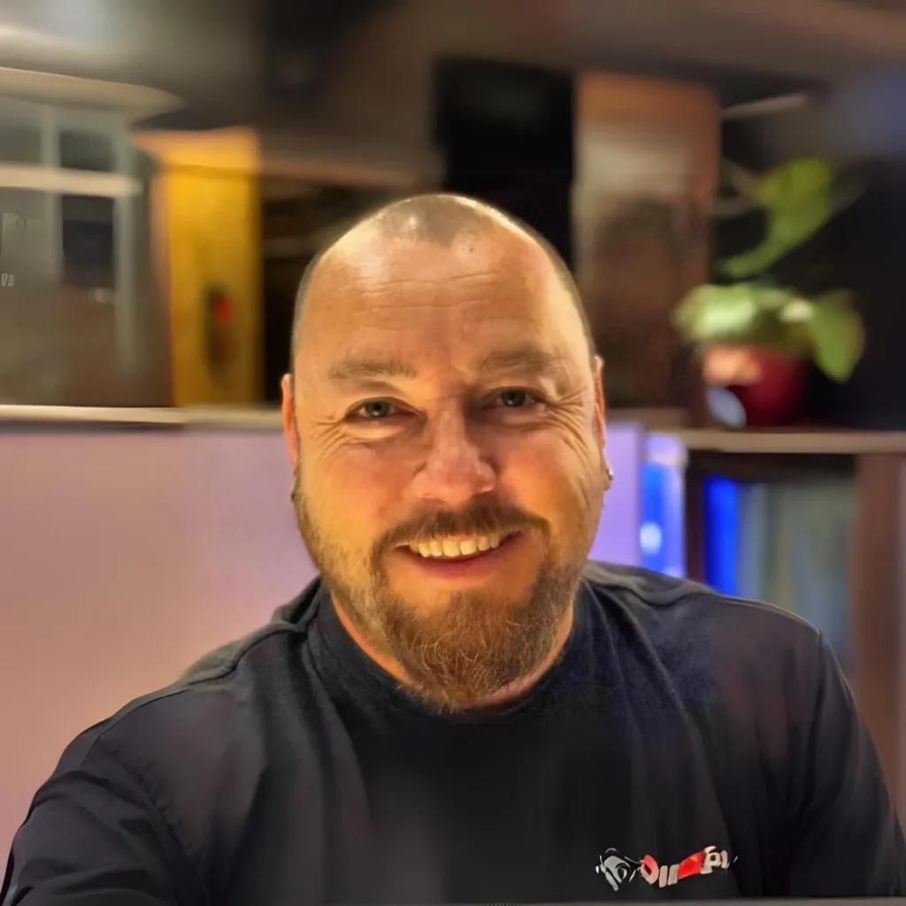 Smiling man with a short beard and shaved head wearing a dark shirt, sitting indoors. Blurred background features a plant and modern decor.