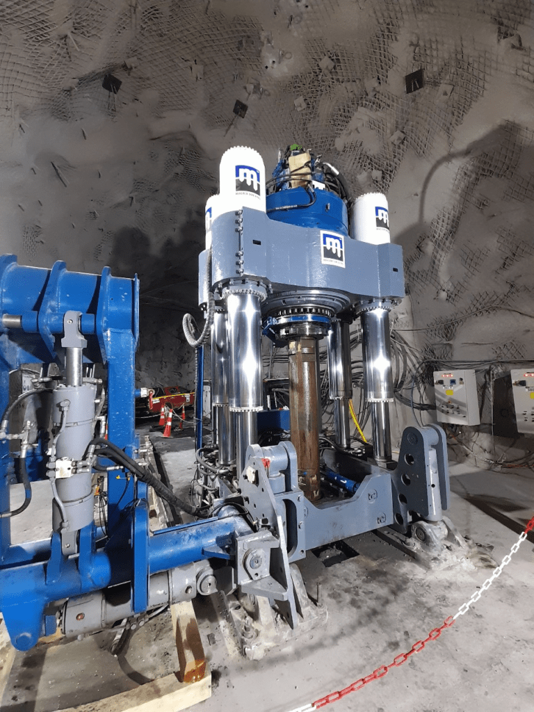 Industrial machinery in a rocky underground setting with grey walls. The equipment is large, predominantly blue and silver, with cylindrical components and multiple connections. The area is lit brightly, revealing cables and additional equipment.