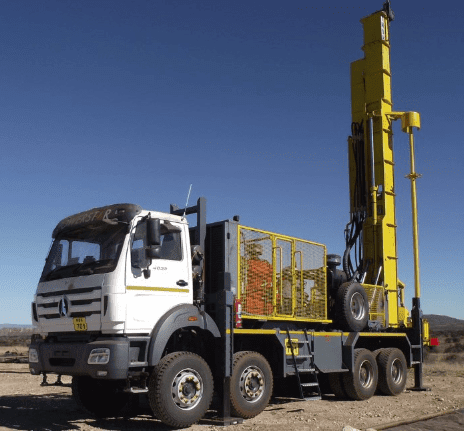 A large drilling truck with a white cab and a tall yellow drilling rig attachment is parked on a barren landscape under a clear blue sky. The truck is equipped with multiple wheels and appears ready for drilling operations.