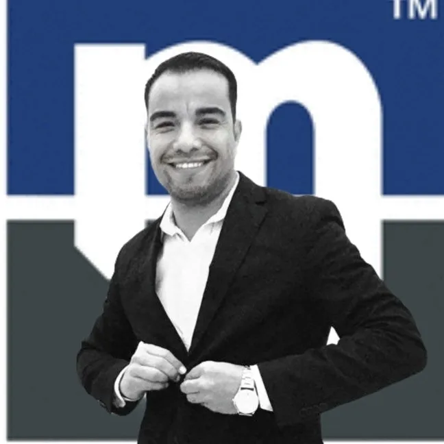 A smiling person in a suit stands in front of a large logo with blue and white colors. The person is buttoning their jacket and wearing a wristwatch. The background features an "m" prominently.