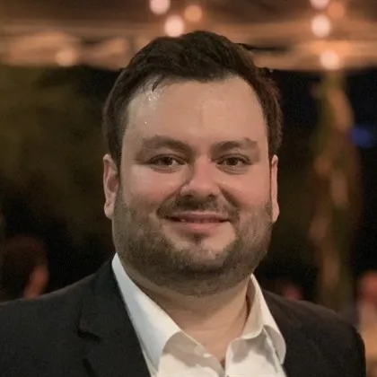 A man with short dark hair and a beard is smiling while wearing a dark suit and white shirt. The background is dimly lit, suggesting an outdoor evening setting with blurred lights.
