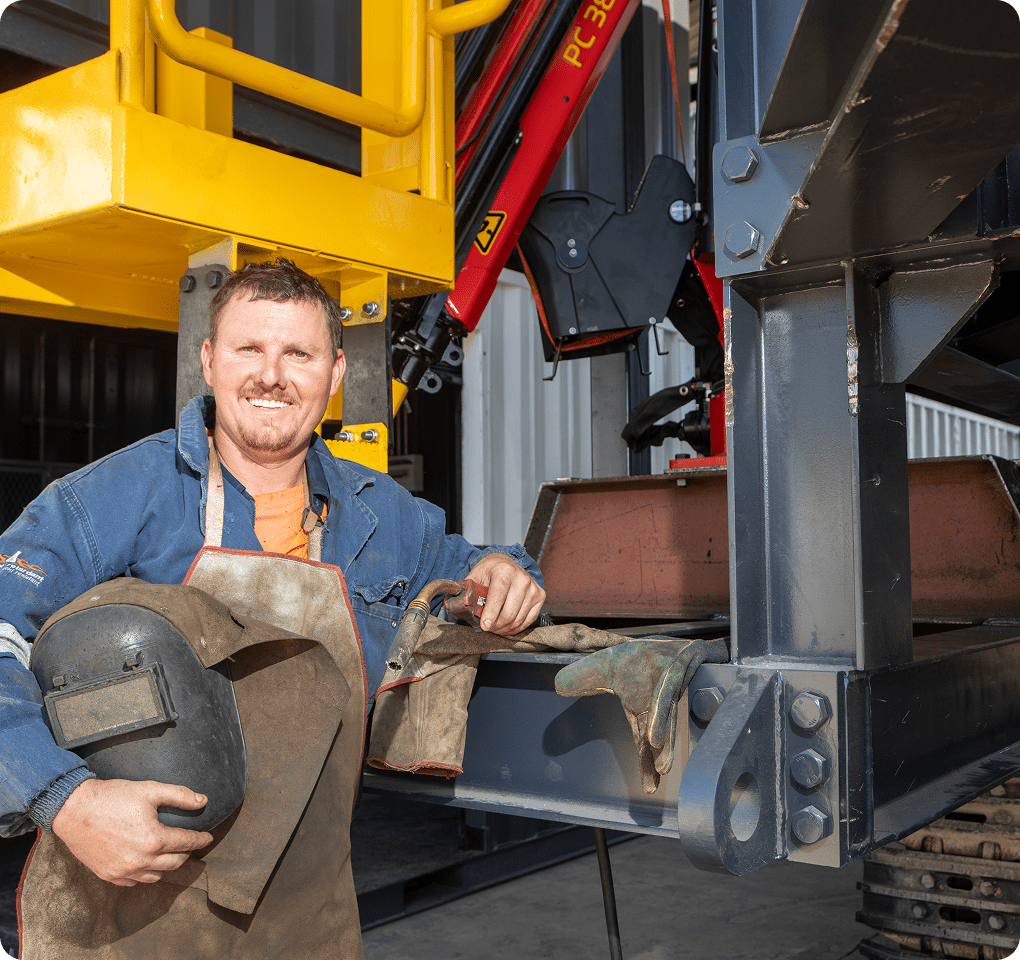 A smiling worker in a blue jacket stands beside industrial machinery, holding a welding mask. He's resting his hand on a large metal structure and wearing heavy-duty gloves. Bright yellow equipment is visible in the background.