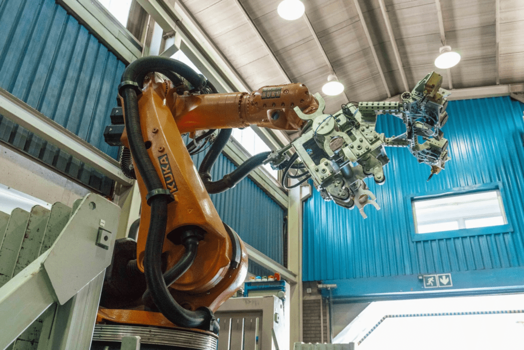 A large industrial robotic arm with a complex metal gripper is positioned inside a spacious, well-lit warehouse. The walls and doors are blue, and overhead lights are visible on the high ceiling.