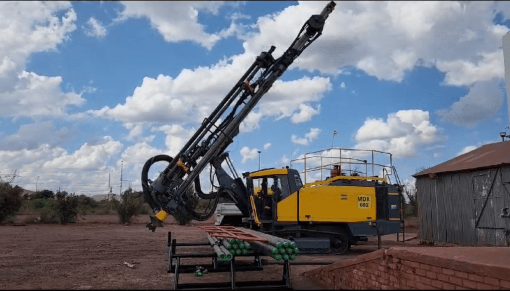 A yellow and black drilling machine is parked outdoors under a partly cloudy sky. Its long arm is raised at an angle. Nearby, a stack of green gas cylinders and a metal platform are visible. A corrugated metal building is in the background.