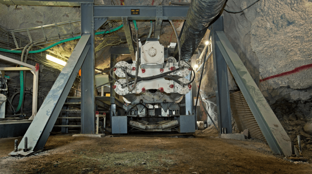 A large piece of industrial machinery is installed inside a rocky tunnel, supported by sturdy metal beams. Various cables and pipes are connected to the machine, indicating an underground setting, such as a mine or a construction site.