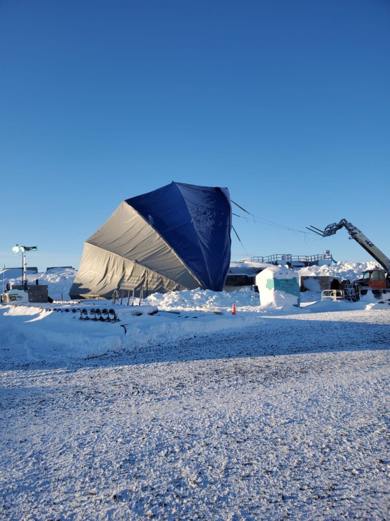 A large blue and gray tarp covers an object amid snowy ground, with equipment, vehicles, and a crane arm nearby under a clear blue sky. Snowbanks and industrial structures are visible in the background.