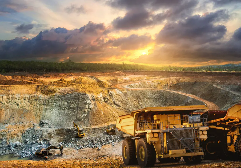 Large mining trucks and excavators operate in an open-pit mine at sunset, with dramatic clouds in the sky and a winding dirt road leading through layers of exposed earth and rock.