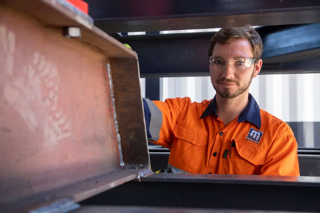 A man wearing safety glasses and an orange high-visibility work shirt stands beside industrial metal beams, looking at the camera, in a workshop or construction setting.
