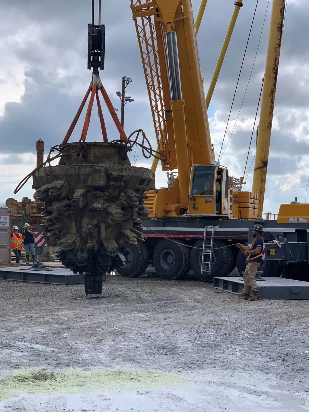 A large yellow crane lifts a massive, cylindrical drilling bit at a construction site while workers in safety gear stand nearby, guiding the operation under a cloudy sky.