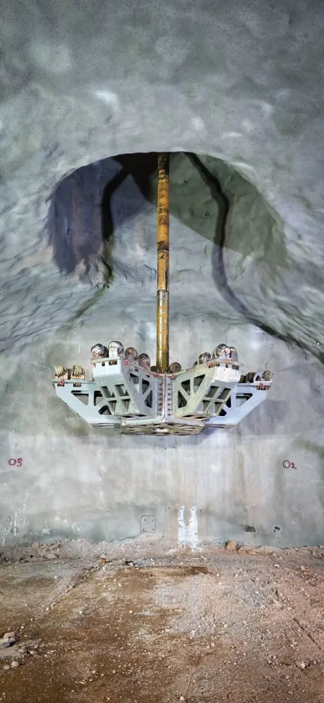 A large tunneling machine suspended from the ceiling of a rocky underground cavern, with its circular cutting head visible and scattered debris on the ground below.